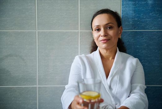 Beautiful positive Hispanic woman wearing white bathrobe, enjoys clean mineral lemon water from glass for body refreshment and following healthy lifestyle. Diet, vitality, skin and body care concept