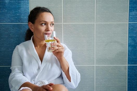 Diet. Vitality. Skin and body care. Healthy habits and lifestyle concept. Beautiful African American woman in white bathrobe, drinking clean lemon water, against bathroom wall background. Copy space