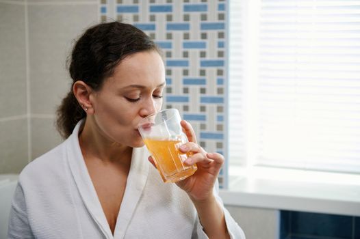 Close-up portrait of a multi-ethnic pretty woman in white bathrobe, drinking water with effervescent antioxidants, minerals, vitamins and nutritional supplements, according to physician's prescription
