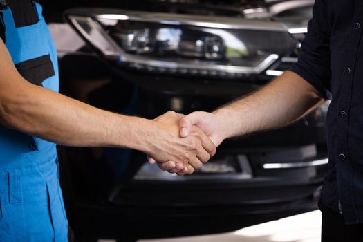 Mechanic and customer shaking hands in an auto repair shop, car service. Two unrecognizable men auto mechanic and client shaking hands at the background of vehicles. Car business, repair services.