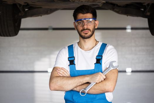 Caucasian bearded man mechanic in uniform with crossed arms and wrench standing at the car repair station. Vehicle service manager worker work in mechanics workshop looking at camera