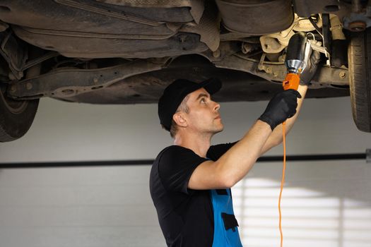 Auto mechanic in blue coveralls and black cap working underneath car lifting machine at the garage. Mechanic check out automobile parts while working with led lamp.