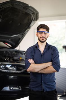 Portrait Bearded Handsome Car Mechanic is Posing in a Car Service. He Wears a Jeans Shirt and Safety Glasses. His Arms are Crossed. Specialist Manager Looks at Camera