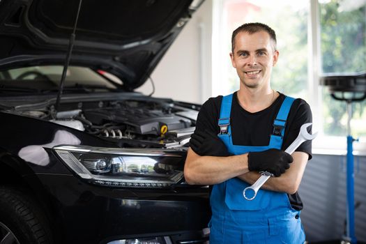 Young caucasian man in blue coveralls holds spanner, smiling and looking into camera. Male car mechanic in spacious repair shop.