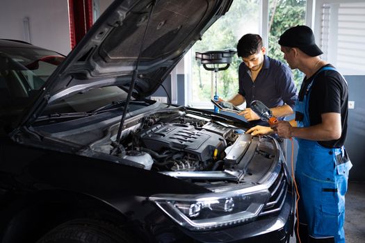 Car service employees inspect car's engine bay with a LED lamp. Manager checks tasts on tablet computer and explains an Engine breakdown to an male mechanic. Auto service.