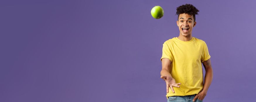 Holidays, vitamins and vacation concept. Portrait of handsome upbeat young male student asking friend something eat, catching apple and smiling happy, standing purple background.