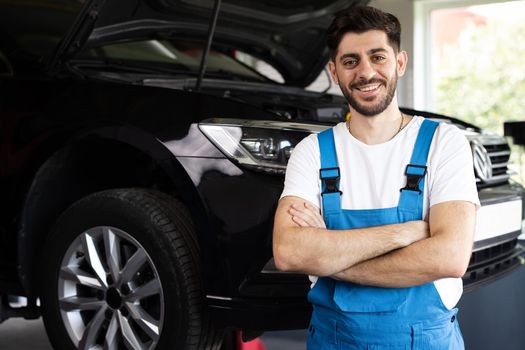Portrait of bearded car mechanic crosses hands in a car workshop in blue uniform with equipment looking into camera. Male car mechanic at workplace in spacious repair shop.