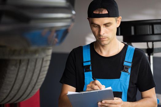 Mechanic inspects the car undercarriage way and makes a note on his inspection sheet. Automobile service, car mechanic. Car service employe inspect car. Modern workshop