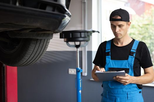 Mechanic inspects the car undercarriage way and makes a note on his inspection sheet. Car service employe inspect car. Automobile service, car mechanic. Modern workshop