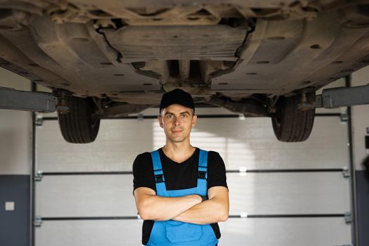 Portrait of mechanic repairing in uniform standing looking camera crossed hands at under lifted car of automobile. Repairing car service concept.