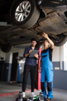 Manager Checks Data on a Notebook and Explains the Breakdown to Mechanic. Car Service Employees Inspect the Bottom of the Car. Workshop. Auto Service.