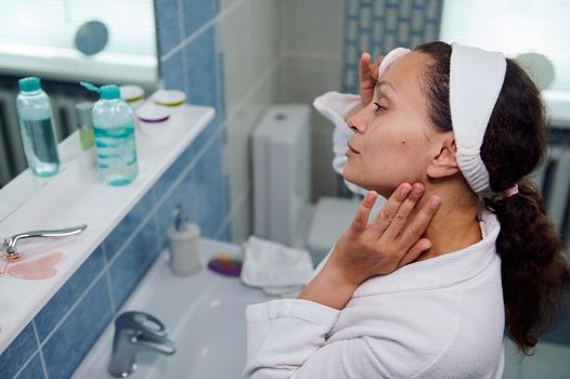 Close-up. Charming curly haired brunette middle-aged woman, applying cleansing lotion, to make her skin clear clean in front of a bathroom mirror, removes cosmetics from her eyes using a cotton pad
