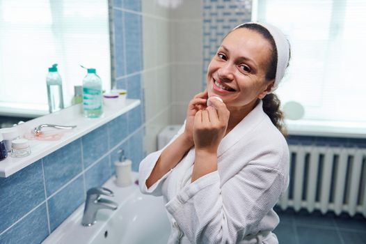 Middle-aged dark-haired charming woman in white bathrobe, using a face cleanser and a cotton pad, removes make-up, and cutely smiles looking at camera standing in the home bathroom. Skin and body care