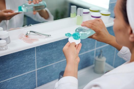 Selective focus on the hands of a blurred woman standing in front of a bathroom mirror and pouring a cleansing lotion on a cotton pad, to clean the face skin and remove make-up. Skin and body care