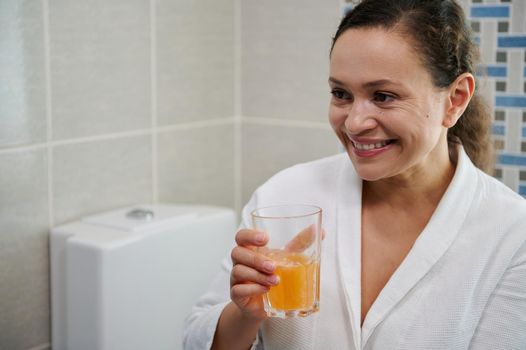 Middle-aged woman in white bathrobe holding a glass of pure water with effervescent tablet, smiling a toothy smile, taking daily dose of vitamins, minerals and dietary nutritional supplement medicine