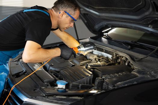 Caucasian male mechanic in blue uniform checking motor in auto service with light. Young man engineer fixing engine. Maintaining concept. Repairment work of technician.