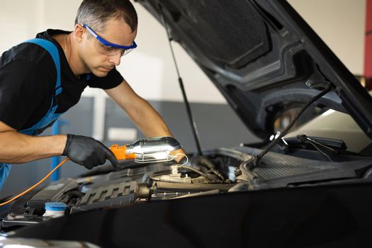 Caucasian Mechanic in Blue Overalls is Working on a Car in a Car Service. Repairman in Safety Glasses is Working on an Usual Car Maintenance. He Hangs Led Lamp. Modern Workshop.