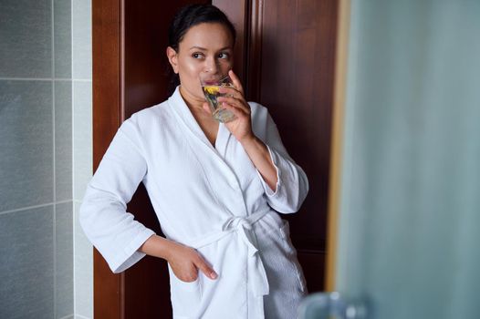 Beautiful African American woman in a white waffle bathrobe drinks refreshing lemon water for healthy and glowing skin. Detox treatment for a good start to the day. Healthy lifestyle. Body care