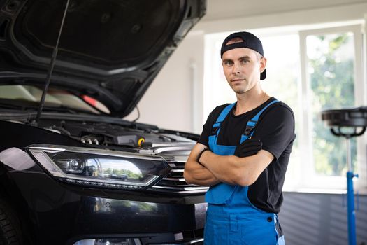 Young Caucasian man in blue coveralls and black cap crosses arms, smiling and looking into camera. Male car mechanic at workplace in spacious repair shop.