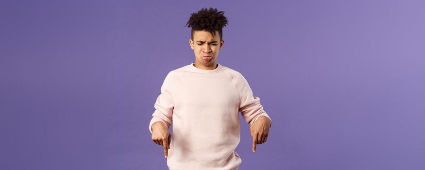 Portrait of gloomy and silly disappointed whining guy, sobbing and crying from envy or jealousy, regret missing chance, looking and pointing down with upset unhappy expression, purple background.