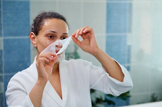 Selective focus on beautiful relaxed brunette woman, reflecting in the bathroom mirror removing a moisturizing smoothing tissue mask from her face. Beauty treatments. Home spa for healthy glowing skin