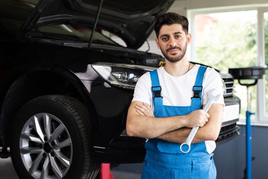 Confident handsome bearded mechanic in blue uniform standing with his arms crossed while workmate working on background. Car mechanic working at automotive service center.