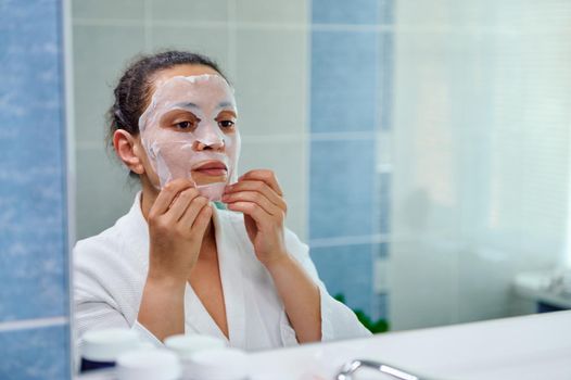 Selective focus on charming brunette woman looking at the bathroom mirror while applying moisturizing smoothing tissue mask on her face. Face care, beauty treatments. Home spa for healthy glowing skin