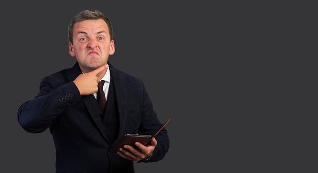 Portrait of a handsome angry man in a blue jacket with a tie holds a gadget in his hand and with the other hand makes a gesture in front of his throat. Negative man on a dark gray background.