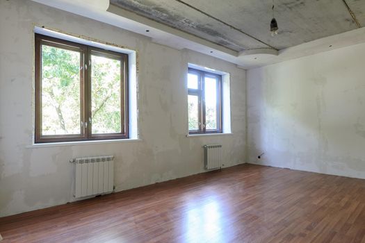 The interior of an empty room during renovation, there are two large windows in the room, heating radiators are located under the windows a