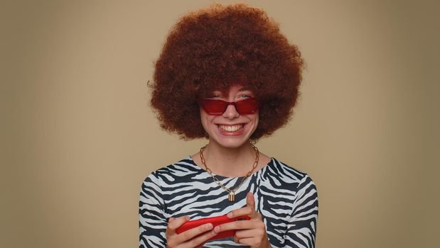 Worried funny young woman with brown lush wig enthusiastically playing racing or shooter video games on smartphone. Adult stylish girl using mobile phone gadget app on beige studio background indoors