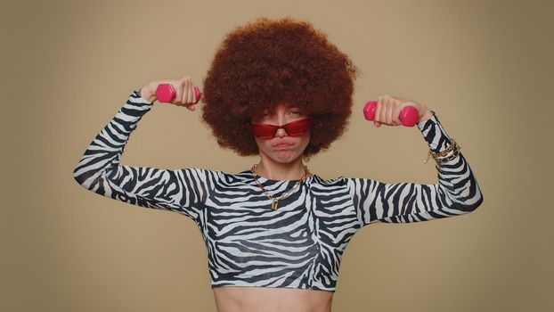 Pretty young woman with brown lush wig working out, pumping up arm muscles lifting pink dumbbells, practicing pilates, healthcare. Adult stylish girl on beige studio background. Sport motivation