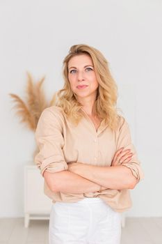 Portrait of a beautiful woman, 30-40 years old, with blond hair, stands in a beige linen shirt and white trousers, in a white room interior. Copy space. Vertical