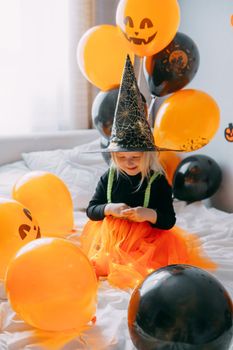 Children's Halloween - a girl in a witch hat and a carnival costume with airy orange and black balloons at home. Ready to celebrate Halloween.