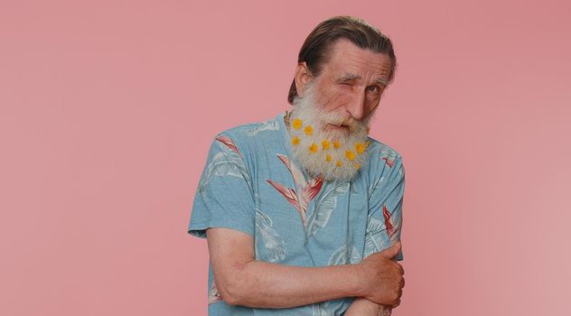 Smiling playful happy senior man with flowers in gray-haired beard blinking eye, looking at camera with toothy smile, winking and flirting, expressing optimism. Elderly grandfather on pink background