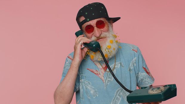 Hey you, call me back. Cheerful bearded old man secretary in t-shirt talking on wired vintage telephone of 60s, asking new client for call. Elderly grandfather isolated on pink studio wall background