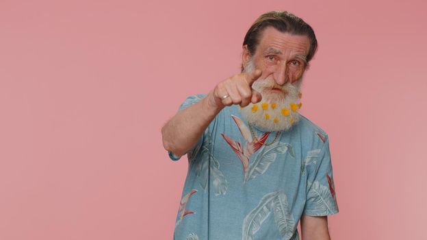 Hey you. Senior man with flowers in beard smiling excitedly and pointing to camera, choosing lucky lottery winner, indicating to awesome you. Elderly grandfather isolated on pink studio background