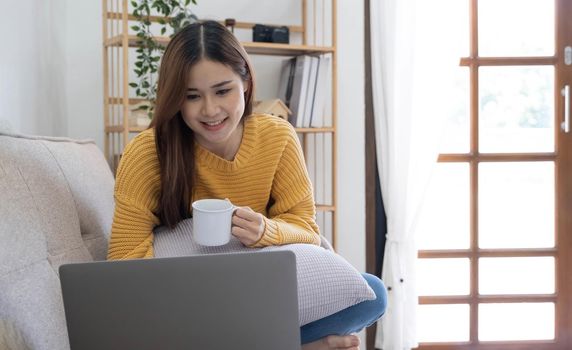 Young asian business woman work from home with laptop computer and drinking coffee on sofa in living room, freelance girl using notebook sitting on couch with comfort and relax, lifestyles concept..