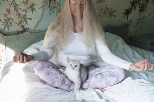 a middle-aged woman with long blond hair meditates in the morning in bed with a kitten,