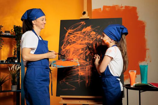 Mother and girl using orange paint on canvas, painting artistic masterpiece with watercolor aquarelle, wet paint from palette and brush. Creating colorful artwork with skills and creative vision.