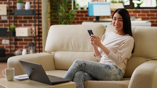 Beautiful woman having smartphone while working remotely and sitting on sofa at home. Attractive person with touchscreen modern device enjoying remote work while relaxing on couch in living room.