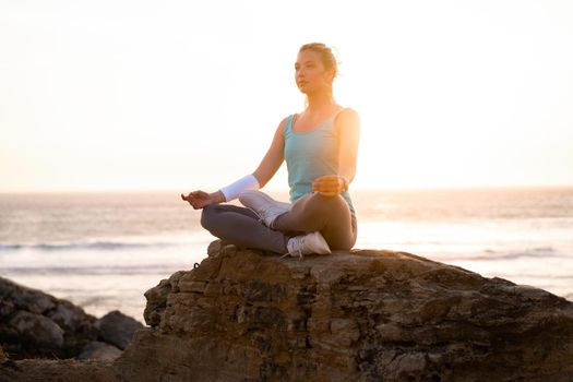 Woman practice yoga lotus pose to meditation with summer vacation rocky beach happiness and relaxation. Calm female exercise with yoga meditate ocean beach with sunset golden time. Zen Mental health