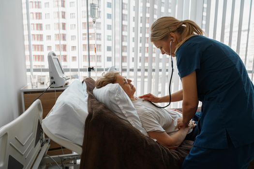 Female doctor listening to old woman's patient breathing, using stethoscope in hospital ward