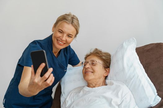 Smiling nurse caring for an elderly woman with a mobile phone takes a selfie in hospital room