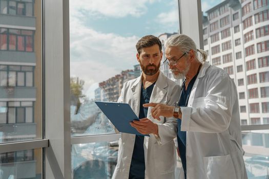 Two doctor colleagues discussing patient diagnosis stand in medicine clinic near panoramic windows