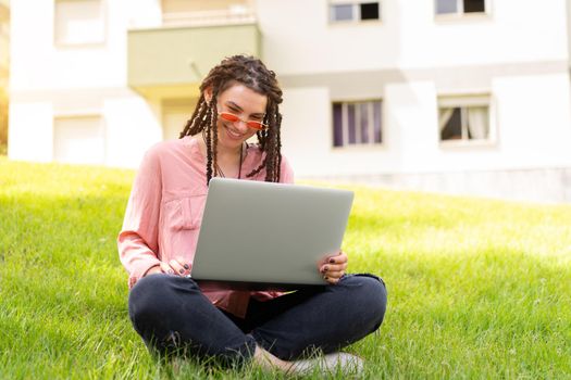 Photo of european woman 25s sitting on green grass in park with legs crossed during summer day while using laptop. Caucasian female hipster with dreadlocks and pink sunglasses on use laptop freelancer