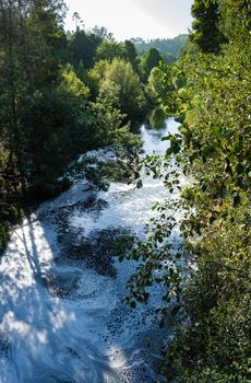 View from the top of the bridge of white foam pollutants swirling across the water surface floating on river Caima, Palmaz - Oliveira de Azemeis - Portugal.