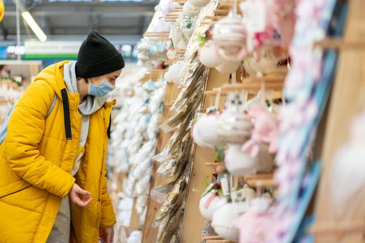 Christmas decoration shopping. Caucasian woman choose new year decor in shopping mall. Medium age female dressed medical mask and yellow winter parka buying Christmas ornament toys indoor store