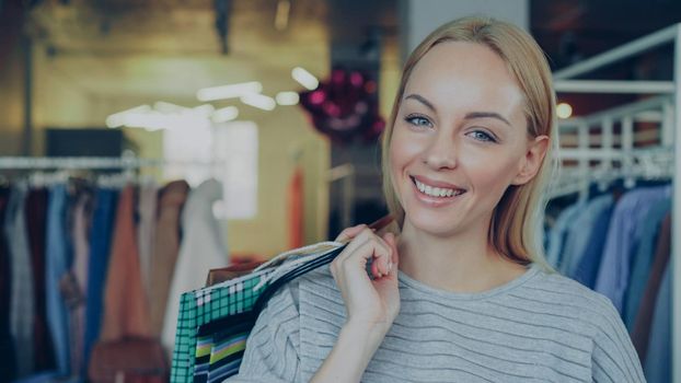 Close-up portrait of cute blond girl standing with paper bags in clothing boutique, moving slightly, laughing and smiling happily and looking at camera. Stylish clothes is in background.