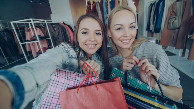 Point of view shot of attractive young ladies making selfie with paper bags in women's clothes boutique. Girls are posing, chatting, and laughing happily. Modern lifestyle concept.