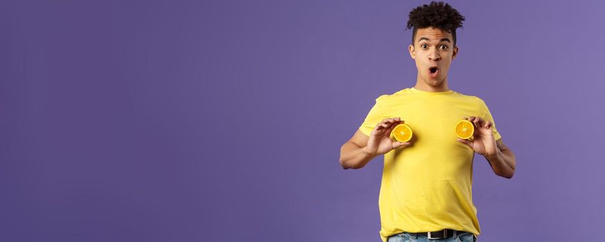 Holidays, vitamins and vacation concept. Portrait of funny and cute young 25s man fool around, showing breast with pieces of oranges over chest, look ashamed or shocked, purple background.